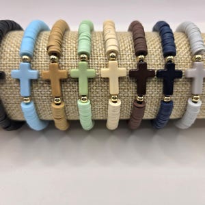 Op de afbeelding: Acht elastische armbanden gemaakt van platte, ronde polymeerklei kralen in zwart, lichtblauw, beige, mintgroen, cr&egrave;me, bruin, donkerblauw en grijs. Elke armband heeft een centrale kruisvormige kraal geflankeerd door twee kleine, ronde, gouden kralen.