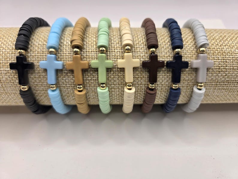 Op de afbeelding: Acht elastische armbanden gemaakt van platte, ronde polymeerklei kralen in zwart, lichtblauw, beige, mintgroen, cr&egrave;me, bruin, donkerblauw en grijs. Elke armband heeft een centrale kruisvormige kraal geflankeerd door twee kleine, ronde, gouden kralen.