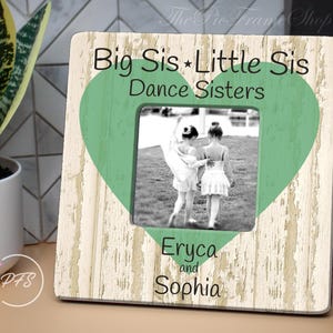 May include: A rustic wooden picture frame with a green heart design. The frame has the text "Big Sis * Little Sis Dance Sisters" and "Eryca and Sophia" printed on it.