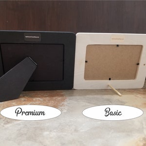 May include: Two picture frames, one black and one white, are shown from the back. The black frame has a black kickstand and the white frame has a wooden kickstand. The black frame is labelled "Premium" and the white frame is labelled "Basic".