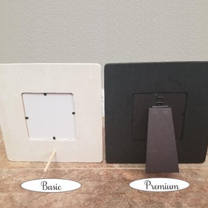 May include: Two picture frames, one white and one black, both with a simple design and a small opening for a photo. The white frame has a wooden stand, while the black frame has a more substantial stand.