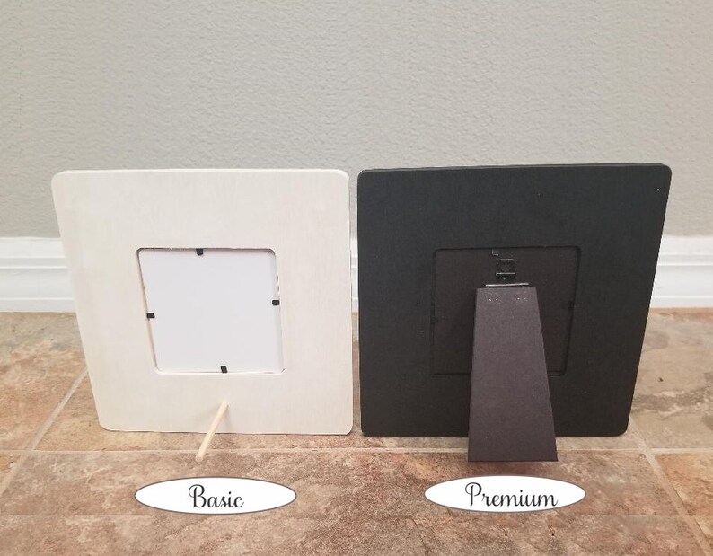May include: Two picture frames, one white and one black, both with a simple design and a small opening for a photo. The white frame has a wooden stand, while the black frame has a more substantial stand.