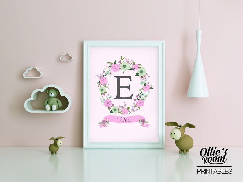 May include: A printable nursery art print with a pink background featuring a floral wreath with the letter E in the center and the name Ella written on a pink ribbon banner below the wreath.