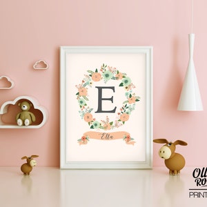 May include: A white framed print with a floral wreath surrounding the letter "E" in gray. The wreath is in shades of peach, green, and white. A peach colored banner below the wreath reads "Ella".