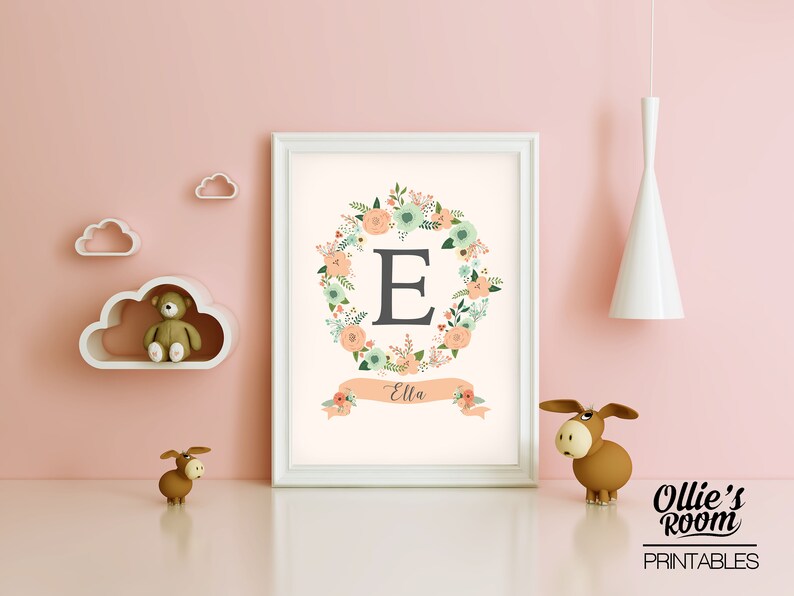 May include: A white framed print with a floral wreath surrounding the letter "E" in gray. The wreath is in shades of peach, green, and white. A peach colored banner below the wreath reads "Ella".