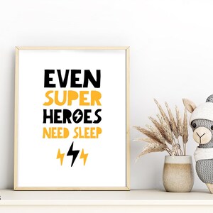 May include: A framed print with a white background and black text that reads "Even Super Heroes Need Sleep" with two yellow lightning bolts underneath. The print is in a light wood frame and is sitting on a white shelf.