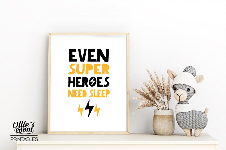 May include: A framed print with a white background and black text that reads "Even Super Heroes Need Sleep" with two yellow lightning bolts underneath. The print is in a light wood frame and is sitting on a white shelf.