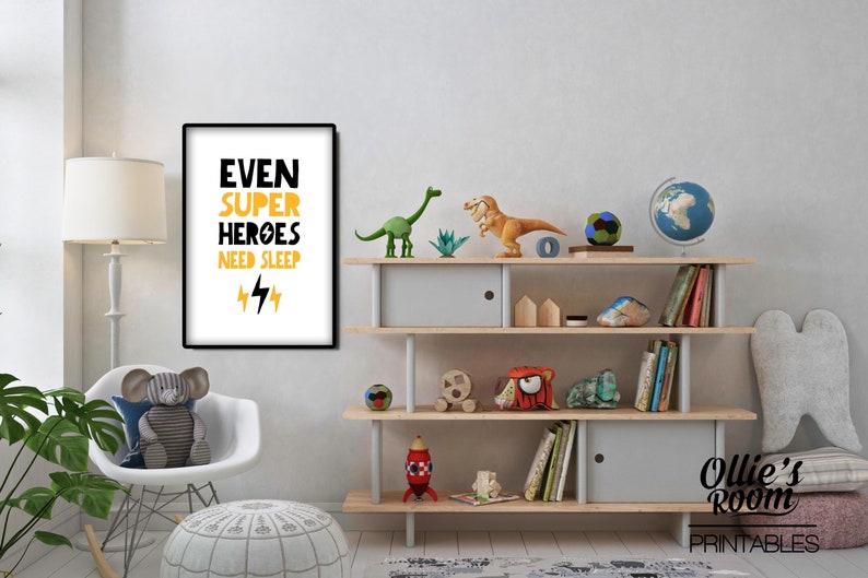 May include: A white framed poster with black and yellow text that reads "Even Super Heroes Need Sleep" with two yellow lightning bolts underneath. The poster is hanging on a white wall above a wooden shelf with a globe, dinosaur toys, and books.