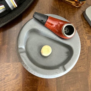 May include: A gray concrete pipe tobacco ashtray with a red and black pipe resting on the edge. The ashtray has a small, round, light brown, wooden tamper in the center.