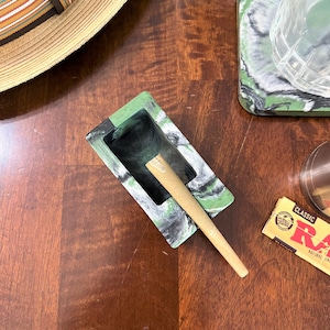 May include: A green and black marbled ashtray with a white rim and a rolled paper inside. The ashtray is sitting on a wooden surface.