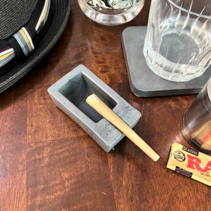 May include: A gray concrete ashtray with a rolled cigarette inside. The ashtray has a rectangular shape with rounded corners and a small indentation for the cigarette. The cigarette is brown and white and is partially sticking out of the ashtray.