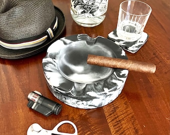 May include: A black and white marbled ashtray with a lit cigar, a black hat, a bottle of tequila, a glass, a coaster, a lighter, and a cigar cutter. The ashtray is round and has a marbled pattern.