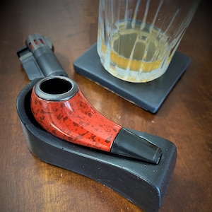 May include: A red and black pipe with a wooden bowl and a black mouthpiece. The pipe is sitting on a black stand.