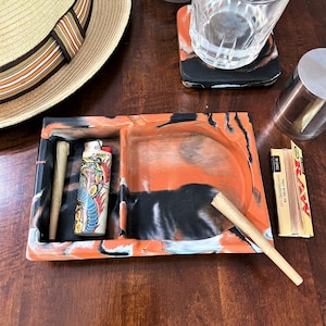 May include: A black and orange tray with two compartments. One compartment contains a lighter with a dragon design and a rolled cigarette. The other compartment has a rolled cigarette. The tray is on a wooden surface.