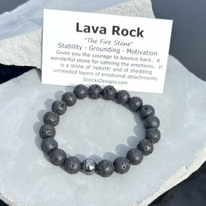 Op de afbeelding: Een zwarte armband van lavastenen kralen met een enkele zilveren kraal. De armband ligt op een grijze achtergrond met een kaart waarop staat: "Lava Rock "The Fire Stone" Stability. Grounding. Motivation Gives you the courage to bounce back. A wonderful stone for calming the emotions. It is a stone of 'rebirth' and of shedding unneeded layers of emotional attachments. StorcksDesigns.com"