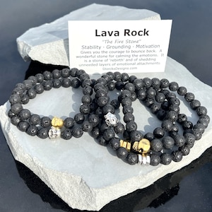Op de afbeelding: Verschillende zwarte lavasteen kralenarmbanden met gouden en zilveren accenten worden gepresenteerd op een wit stenen oppervlak. Een kaart met de tekst "Lava Rock" en productdetails is zichtbaar.