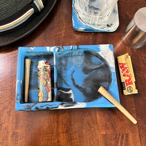 May include: A blue and white marble rolling tray with three compartments. The tray has a round compartment in the center, a rectangular compartment on the left, and a smaller rectangular compartment on the right. A lighter and two rolling papers are on the tray.