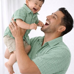 Puede incluir: Un hombre sostiene a un bebé riendo en el aire. Ambos visten camisas verde claro. El bebé también lleva pantalones cortos color caqui. El hombre sonríe con la boca abierta y el bebé ríe con la boca abierta. Momento feliz.