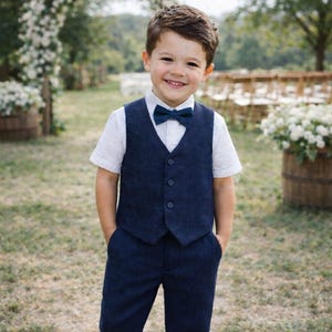 May include: A young boy in a navy blue vest, pants, and bow tie, paired with a white shirt and brown shoes. The boy is smiling and standing outdoors in front of a floral arch and wooden barrels.