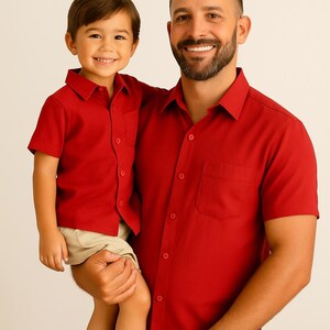 May include: A father holding a child, both wearing matching red short-sleeved shirts with a chest pocket and button-down collar. The child wears khaki shorts. The father is smiling.