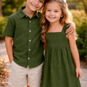 Pode incluir: Um menino e uma menina sorrindo, abraçando-se. O menino usa uma camisa verde-oliva de manga curta com botões e calções bege. A menina usa um vestido verde-oliva com decote quadrado. O fundo está desfocado com luz natural.