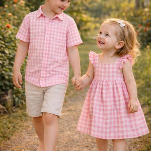 May include: A boy and a girl walk together, holding hands. The boy is wearing a pink and white checked shirt and beige shorts. The girl wears a pink and white checked dress. Both children are barefoot, walking on a path.
