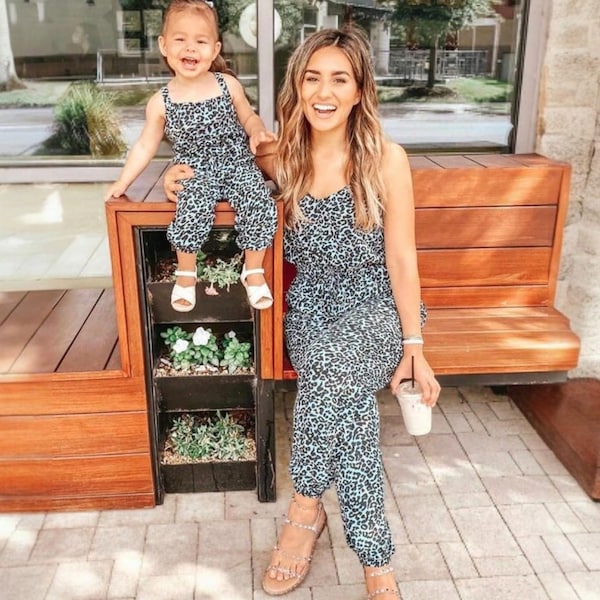 May include: A woman and a young girl wearing matching blue and white animal print jumpsuits. The woman is wearing sandals and holding a beverage. The girl is wearing white sandals. They are sitting on a wooden bench.