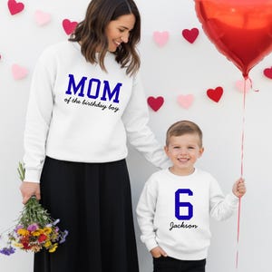 May include: A woman and a young boy wearing matching white sweatshirts. The woman's sweatshirt says "MOM of the birthday boy" in blue. The boy's sweatshirt has a blue "6" and the name "Jackson". A red heart-shaped balloon is held by the boy.