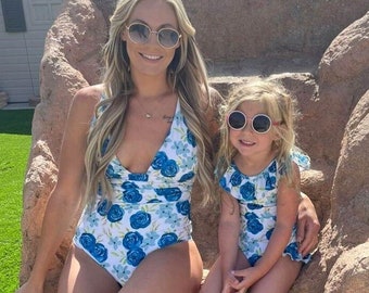 May include: A mother and daughter wearing matching blue and white floral print swimsuits. The woman is wearing sunglasses and the girl is wearing round sunglasses. They are sitting on a large rock.