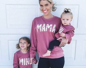 May include: Three people wearing matching pink sweatshirts with floral print lettering. The adult is wearing black leggings and holding a child in their arms. The child in the adult's arms is wearing a pink sweatshirt with the word "Mini" on it. The other child is wearing a pink sweatshirt with the word "Mini" on it and black leggings.