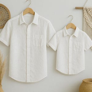 May include: Two white linen short-sleeve shirts hanging on wooden hangers. The shirts have a button-down front, a chest pocket, and a collar. The shirts are displayed against a neutral background, suggesting a minimalist aesthetic.