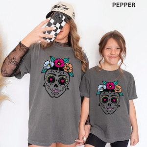 Könnte beinhalten: Zwei anthrazitfarbene T-Shirts mit einer Grafik eines schwarzen Zuckerschädels, der mit einer Krone aus blauen, rosa und orangefarbenen Blumen verziert ist. Der Schädel hat herzförmige Augen und einen genähten Mund. Der Text PEPPER ist oben rechts.