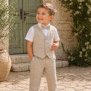 Puede incluir: Un niño pequeño con un traje de lino beige de tres piezas: camisa blanca de manga corta, chaleco y pantalón a juego. El conjunto incluye una pajarita beige y zapatos de ante marrón. El niño sonríe y está de pie al aire libre.