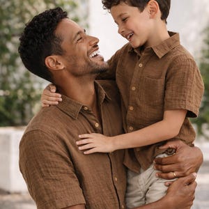 May include: A man and a child are embracing and smiling. Both are wearing matching short-sleeved, button-down shirts in a brown color. The child is wearing khaki shorts. The background is blurred, suggesting an outdoor setting.