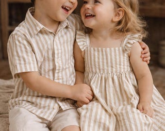 Puede incluir: Un niño y una niña sonríen y se abrazan. El niño lleva una camisa a rayas beige y blanca y pantalones cortos. La niña lleva un vestido a rayas a juego con mangas con volantes. Ambos llevan sandalias marrones. La ropa es de tela ligera.