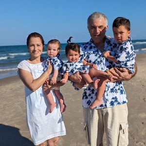 K&ouml;nnte beinhalten: Eine vierk&ouml;pfige Familie am Strand, die Eltern halten zwei Babys, alle tragen passende blaue und wei&szlig;e Hemden mit tropischem Muster. Die Eltern tragen Khaki-Shorts und die Mutter ein wei&szlig;es Kleid.