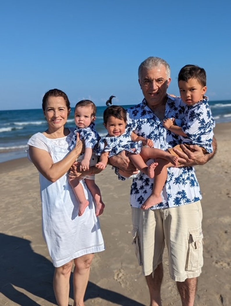K&ouml;nnte beinhalten: Eine vierk&ouml;pfige Familie am Strand, die Eltern halten zwei Babys, alle tragen passende blaue und wei&szlig;e Hemden mit tropischem Muster. Die Eltern tragen Khaki-Shorts und die Mutter ein wei&szlig;es Kleid.