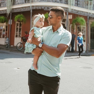 Puede incluir: Un hombre con gafas de sol y una camisa verde claro sostiene a un beb&eacute; con un vestido y una diadema verde claro a juego. El beb&eacute; mira al hombre. El hombre lleva un reloj y pantalones cortos negros.