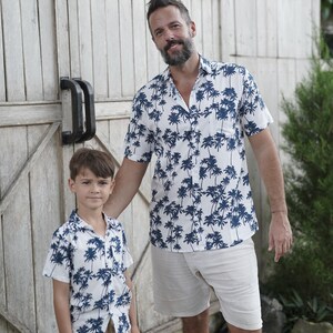 K&ouml;nnte beinhalten: Ein Vater und sein Sohn tragen passende wei&szlig;e Hemden mit kurzen &Auml;rmeln und einem blauen Palmenprint. Die Hemden haben einen Knopfkrag und kurze &Auml;rmel. Der Vater tr&auml;gt beige Shorts und der Sohn tr&auml;gt Khaki-Shorts.