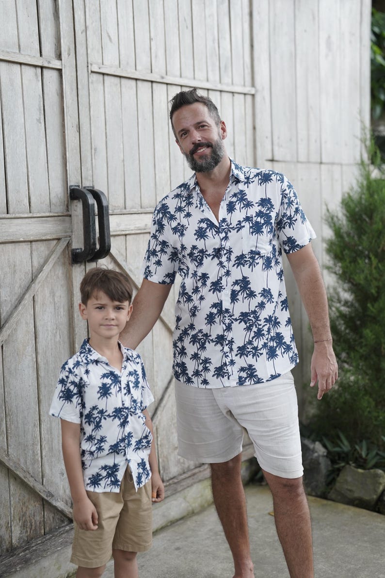 K&ouml;nnte beinhalten: Ein Vater und sein Sohn tragen passende wei&szlig;e Hemden mit kurzen &Auml;rmeln und einem blauen Palmenprint. Die Hemden haben einen Knopfkrag und kurze &Auml;rmel. Der Vater tr&auml;gt beige Shorts und der Sohn tr&auml;gt Khaki-Shorts.