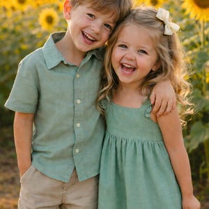 Pode incluir: Um menino e uma menina sorrindo se abraçam em um campo de girassóis. O menino usa uma camisa verde clara e calções cáqui. A menina usa um vestido verde claro e um laço branco. O fundo é preenchido com girassóis.