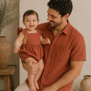 May include: A baby in a rust-coloured romper with ruffle sleeves is held by a person wearing a matching short-sleeved shirt. The person is wearing beige trousers. The background includes a wooden stool and a decorative vase.