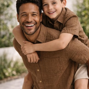 May include: A man and a child are smiling at the camera. Both are wearing brown button-down shirts. The man is carrying the child on his back. The background is blurred with green foliage.