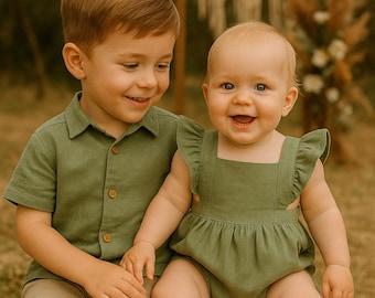 Puede incluir: Dos niños con conjuntos verde oliva. El mayor lleva una camisa abotonada y pantalones cortos color caqui. El más pequeño lleva un pelele con mangas con volantes. Ambos niños sonríen.