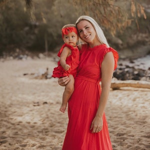 Puede incluir: Una mujer sostiene a un beb&eacute;, ambos con vestidos y diademas rojos a juego. El vestido de la mujer tiene mangas con volantes y una falda fluida. Est&aacute;n de pie en una playa de arena con &aacute;rboles al fondo.