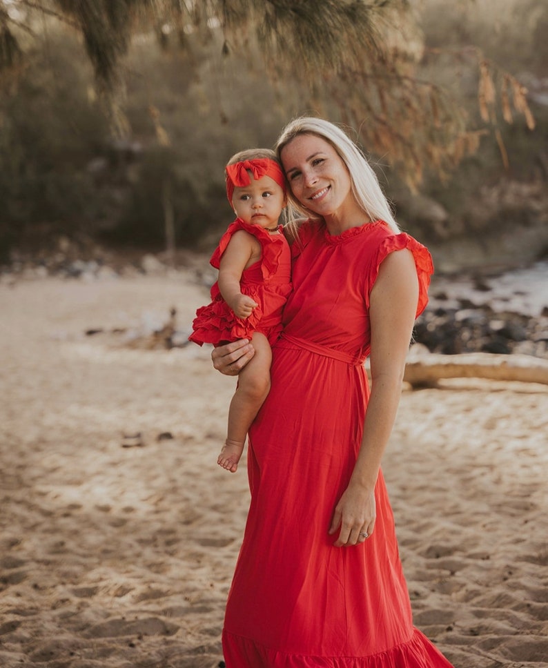 Puede incluir: Una mujer sostiene a un beb&eacute;, ambos con vestidos y diademas rojos a juego. El vestido de la mujer tiene mangas con volantes y una falda fluida. Est&aacute;n de pie en una playa de arena con &aacute;rboles al fondo.