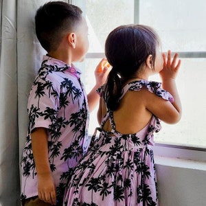 K&ouml;nnte beinhalten: Zwei Kinder in passenden pinkfarbenen und schwarzen Kleidern mit Palmenprint stehen an einem Fenster. Das M&auml;dchen tr&auml;gt ein &auml;rmelloses Kleid und der Junge ein kurz&auml;rmeliges Hemd.