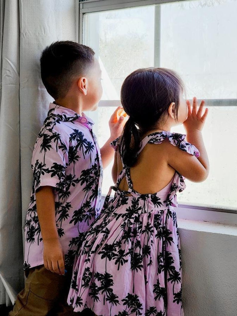 K&ouml;nnte beinhalten: Zwei Kinder in passenden pinkfarbenen und schwarzen Kleidern mit Palmenprint stehen an einem Fenster. Das M&auml;dchen tr&auml;gt ein &auml;rmelloses Kleid und der Junge ein kurz&auml;rmeliges Hemd.