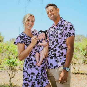 K&ouml;nnte beinhalten: Eine dreik&ouml;pfige Familie tr&auml;gt passende Kleidung mit Palmen-Print in Lila und Schwarz. Der Vater h&auml;lt ein Baby M&auml;dchen im Arm, das ein passendes Outfit und eine schwarze Schleife tr&auml;gt. Die Familie steht vor einem gr&uuml;nen Busch.