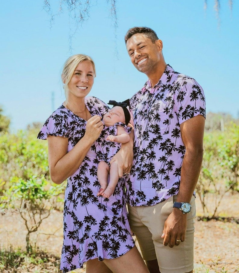 K&ouml;nnte beinhalten: Eine dreik&ouml;pfige Familie tr&auml;gt passende Kleidung mit Palmen-Print in Lila und Schwarz. Der Vater h&auml;lt ein Baby M&auml;dchen im Arm, das ein passendes Outfit und eine schwarze Schleife tr&auml;gt. Die Familie steht vor einem gr&uuml;nen Busch.